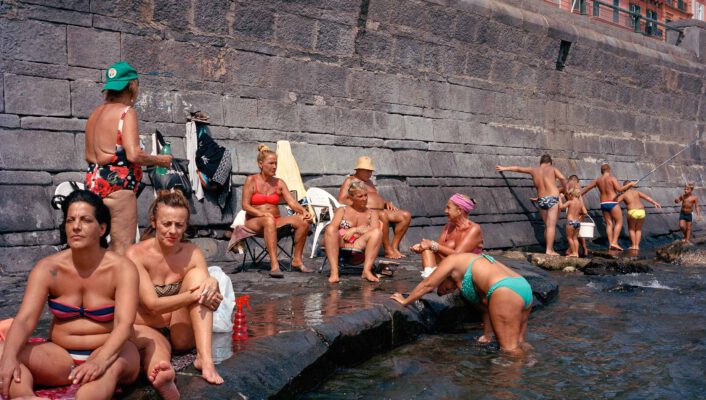 a group of people sitting along the rocky wall against the sea in Naples