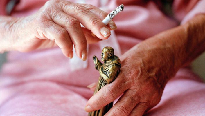 someone in a pink dress holding a small statue of a pope in one hand and a cigarette in the other