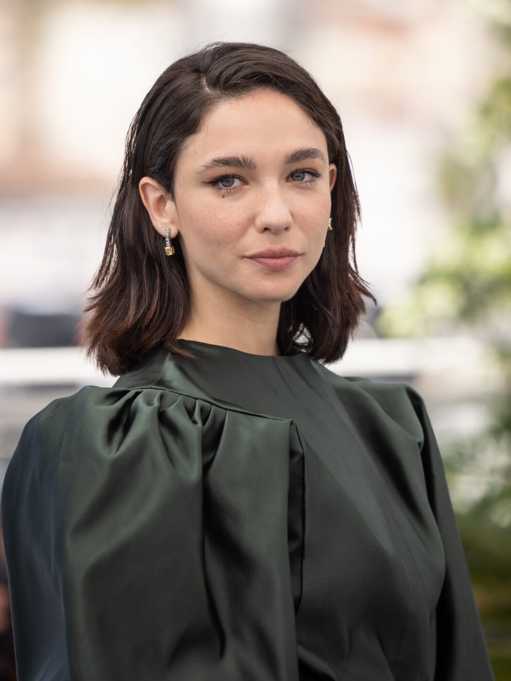 A woman with short dark brown hair, light eyes, and a dark green satin top stands outdoors, gazing neutrally at the camera.