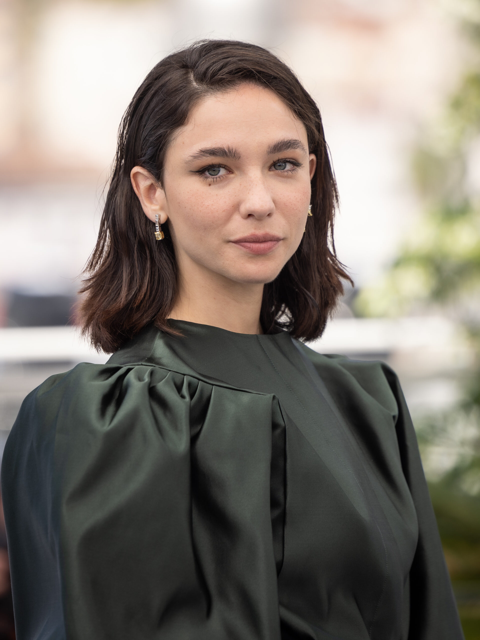A woman with short dark brown hair, light eyes, and a dark green satin top stands outdoors, gazing neutrally at the camera.