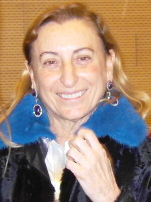 A smiling woman with light brown hair in blue earrings and a black coat with blue fur collar stands before a tan, textured wall.