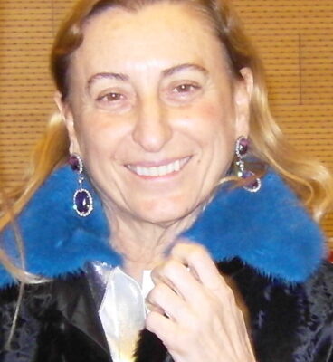 Una donna sorridente dai lunghi capelli castano chiaro, con un cappotto scuro con collo di pelliccia blu e orecchini vistosi, si trova davanti a uno sfondo marrone.