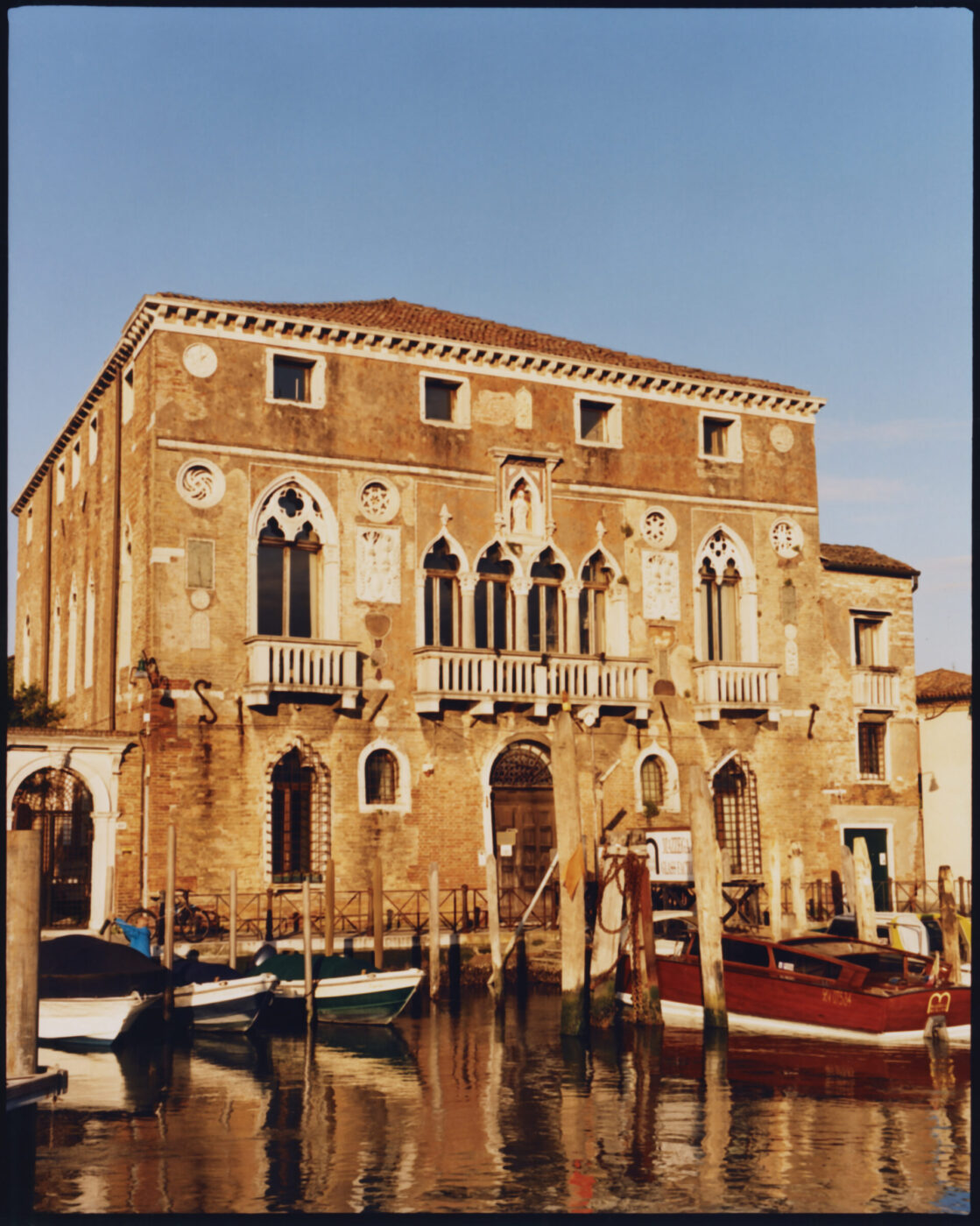 A historic Venetian building with arched windows and balconies stands by a canal in Murano, Venice, with several colorful boats moored along wooden posts in the water under a clear blue sky.