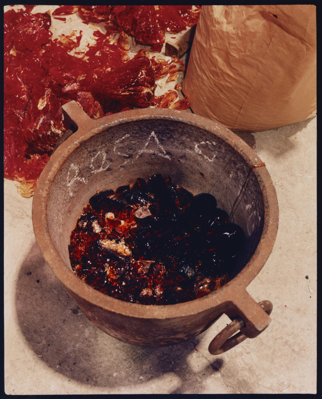 Red Resin Preparation - A large metal pot contains a dark red, melted substance resembling resin or chemicals, reminiscent of Murano Venice glassmaking. Red, translucent solid chunks are scattered in the background near a brown paper bag on a light-colored surface.