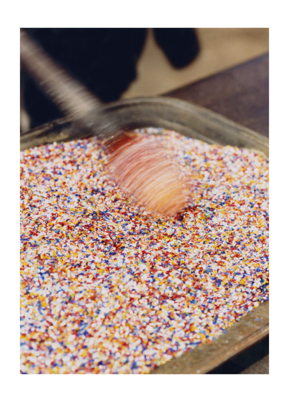 [Una mano mescola un grande vassoio pieno di caramelle colorate con un utensile di legno. Il vibrante mix di rosso, giallo, blu e bianco ricorda l'arte vetraria di Murano Venezia, in quanto la sfocatura fonde insieme le tonalità.