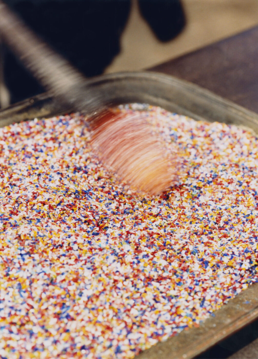 A hand stirring vibrant sprinkles - A hand is stirring a large metal tray filled with colorful sprinkles, creating a blur of motion above the vibrant mixture reminiscent of Murano Venice glass artistry.