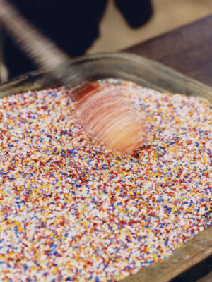 A hand stirring vibrant sprinkles - A hand is stirring a large metal tray filled with colorful sprinkles, creating a blur of motion above the vibrant mixture reminiscent of Murano Venice glass artistry.