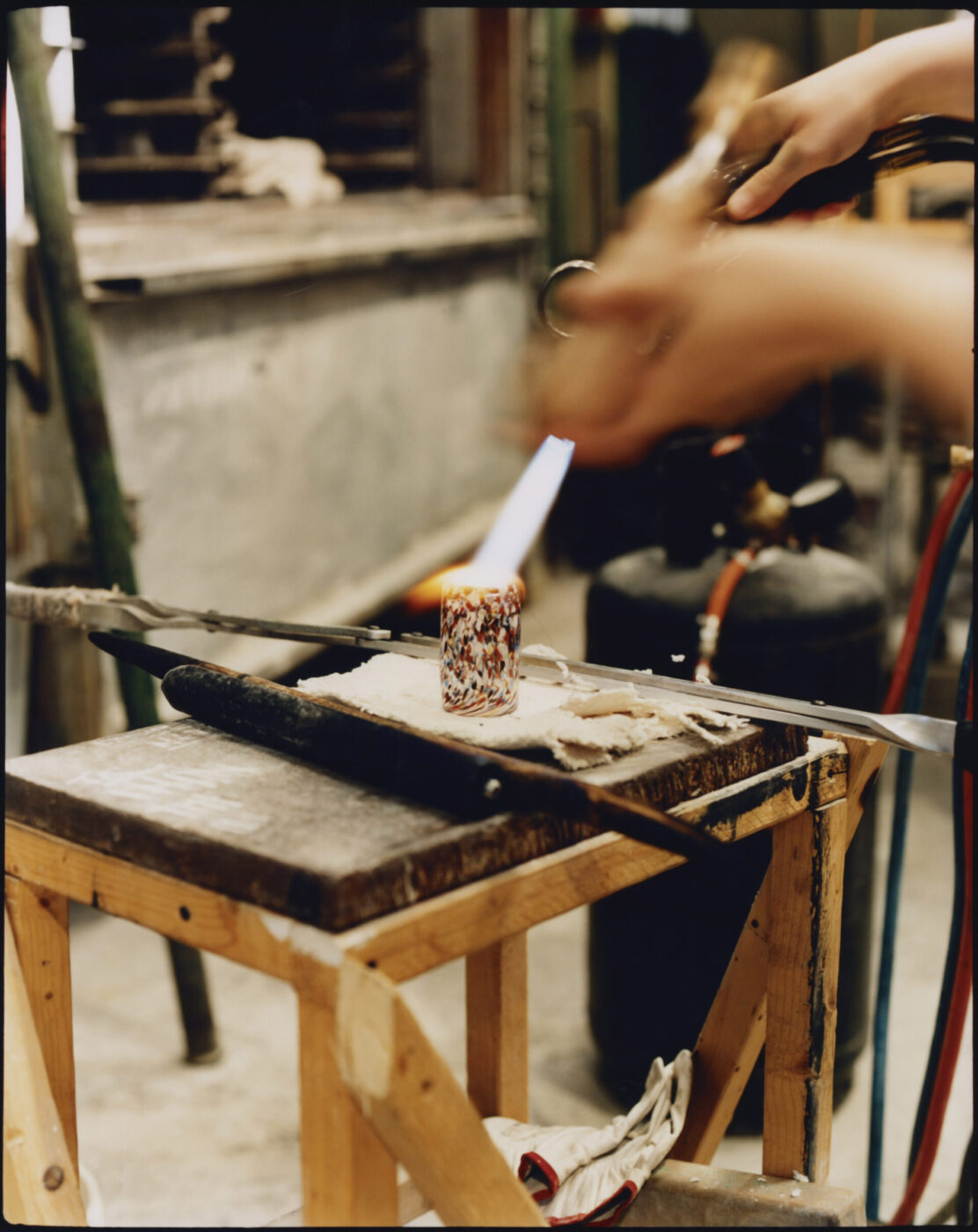 Venetian Glassblowing - A person uses a torch to heat a colorful glass object on a workbench in a Murano Venice-inspired glassblowing studio, with tools and equipment visible in the background.