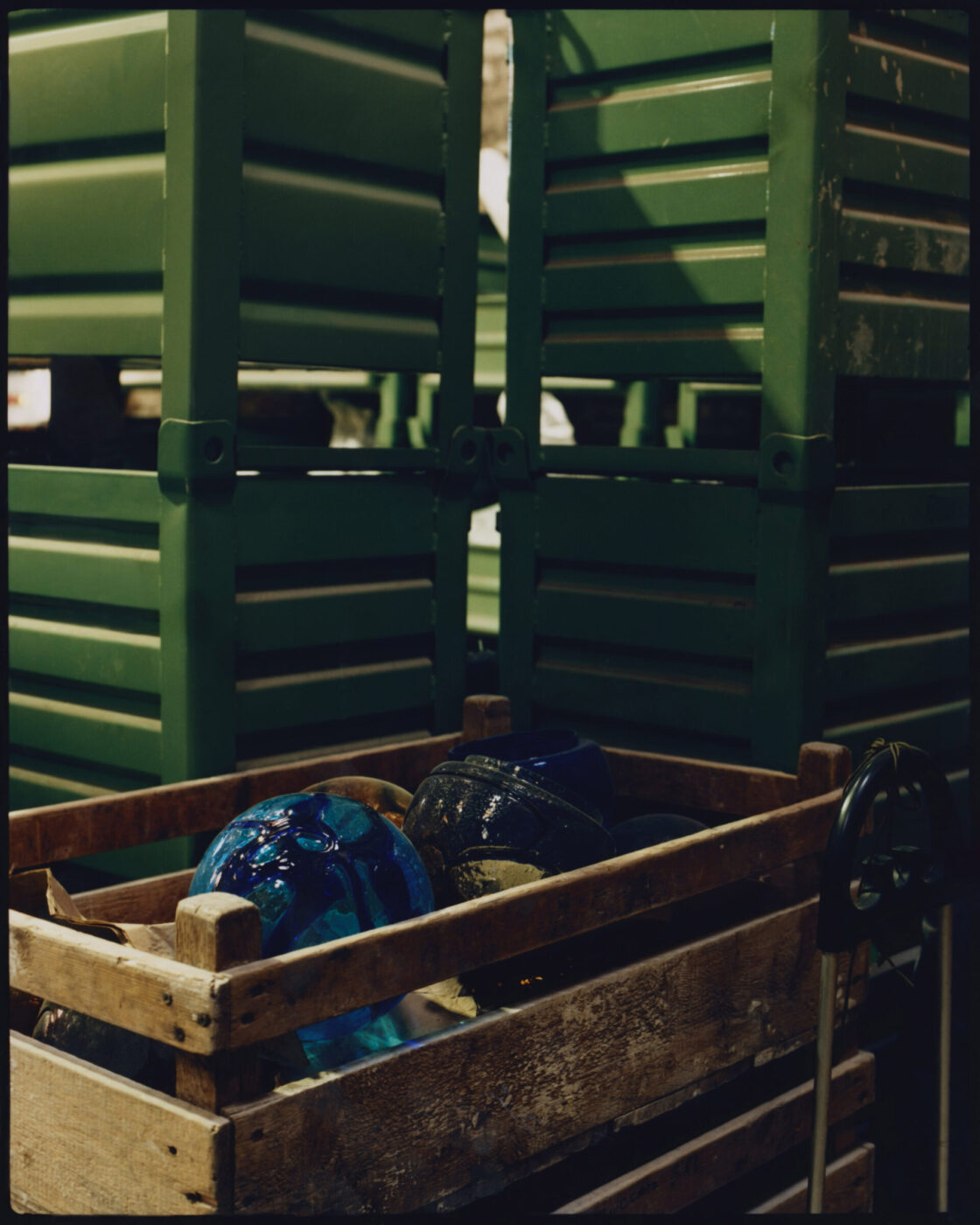A wooden crate containing colorful Murano Venice glass spheres - A wooden crate filled with vibrant, multicolored Murano glass spheres is placed in front of a stack of green metal chairs. The dim lighting casts dramatic shadows and enhances the rich hues, creating a moody atmosphere.