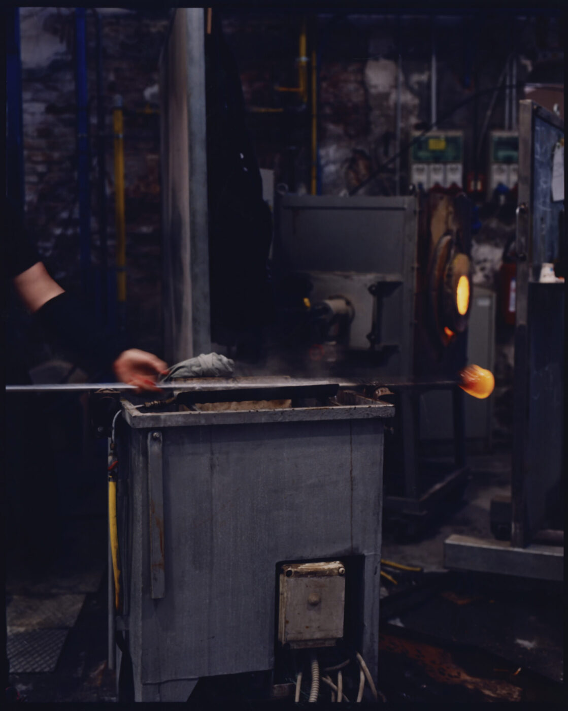 A person shapes hot molten glass on a rod in an industrial workshop reminiscent of Murano Venice, with furnaces glowing in the background and machinery surrounding the workspace. For example, if your image title is Venetian Glassblowing Workshop, then the alt text would be: Venetian Glassblowing Workshop - A person shapes hot molten glass on a rod in an industrial workshop reminiscent of Murano Venice, with furnaces glowing in the background and machinery surrounding the workspace.