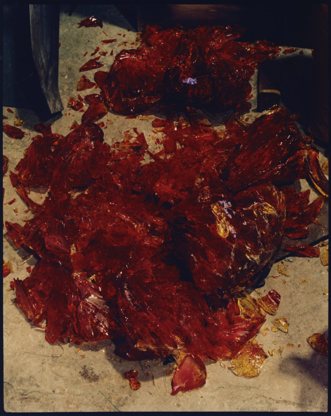 Large, jagged shards of bright red resin reminiscent of Murano Venice glass are scattered across a concrete floor, creating a dramatic, colorful contrast against the neutral surface.