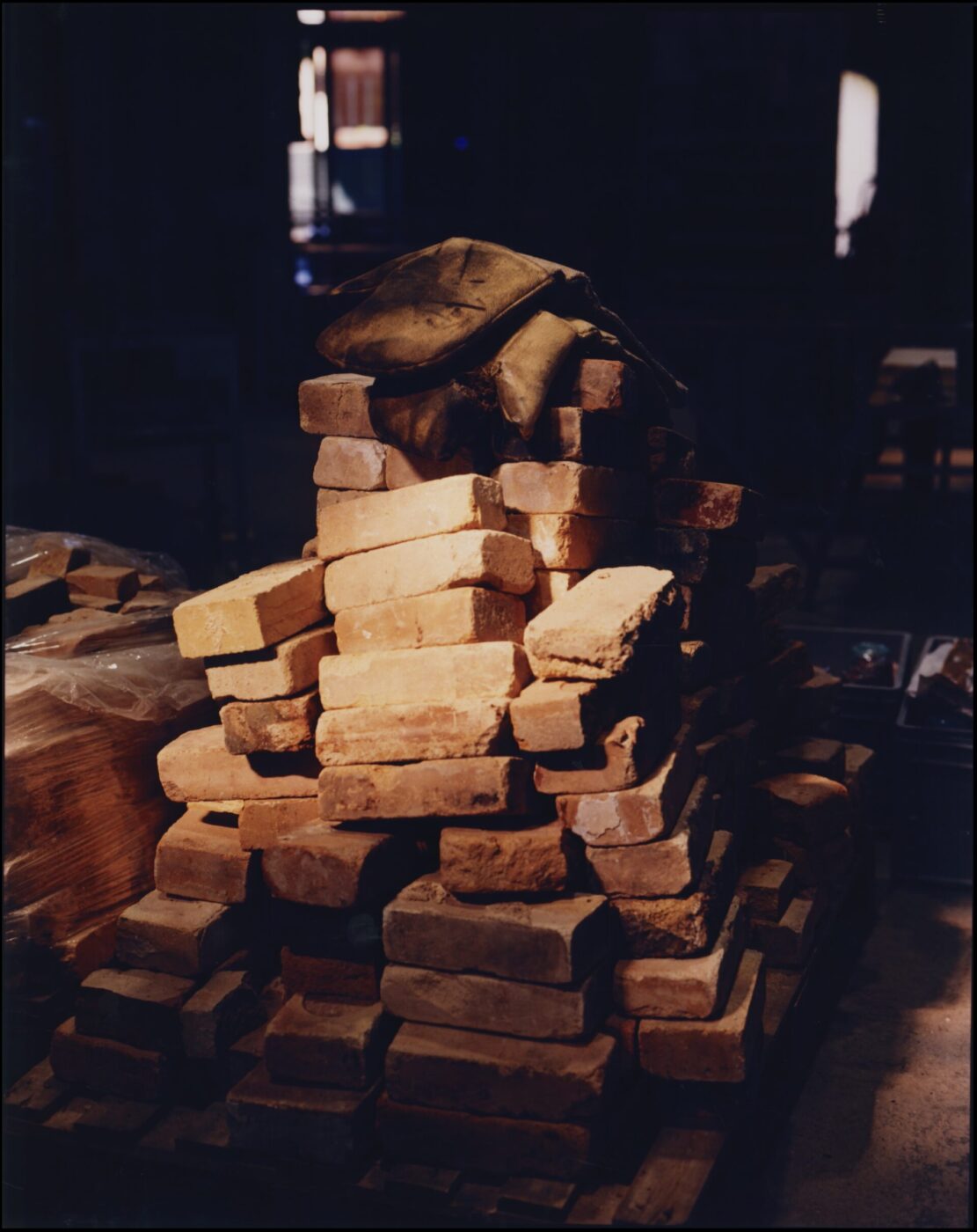 A stack of unevenly piled bricks sits in a dimly lit area. On top rests a worn brown bag, reminiscent of Murano Venice craftsmanship. Light from a doorway or window illuminates part of the scene, casting dramatic shadows. If an image title is given, prepend it to the beginning as described: [Image Title] - A stack of unevenly piled bricks sits in a dimly lit area. On top rests a worn brown bag, reminiscent of Murano Venice craftsmanship. Light from a doorway or window illuminates part of the scene, casting dramatic shadows.