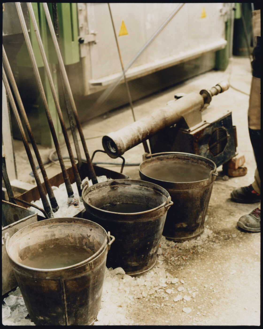 Three metal buckets filled with water sit on a workshop floor scattered with glass shards, evoking the artistry of Murano Venice. Metal rods lean against the wall, industrial equipment appears in the background, and a person’s legs and shoes are partially visible.