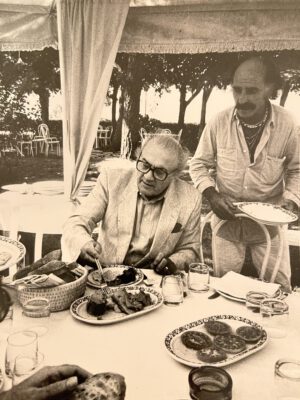 Vintage picture of filmaker federico Fellini formal dress and wearing glasses eating over a tables filled up with italian dishes and ceramic plates