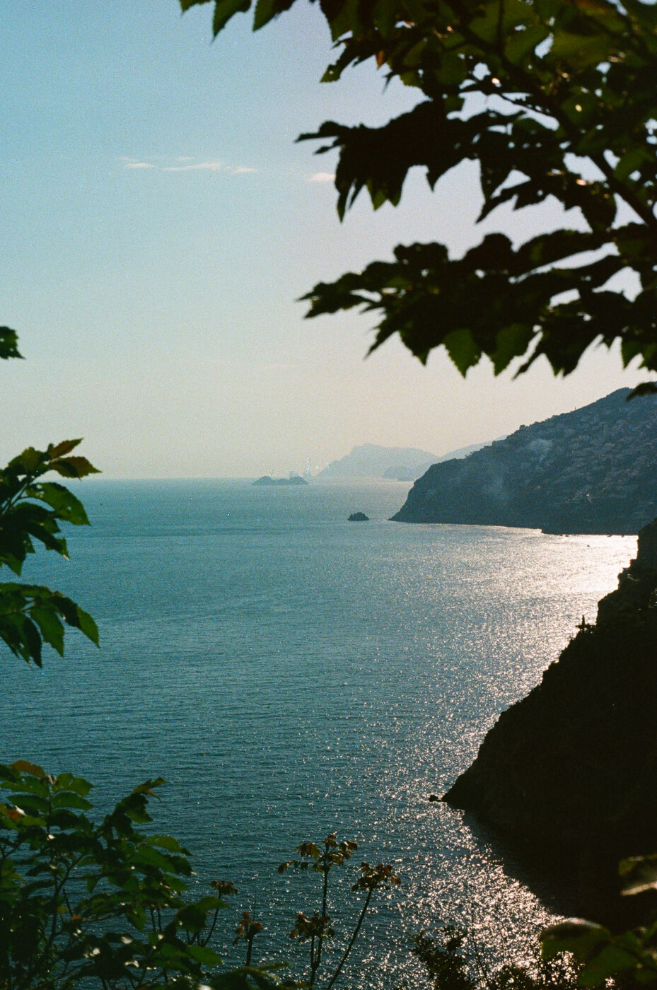 Costiera Amalfitana - Veduta di un mare calmo e blu scintillante delimitato da scogliere frastagliate sulla Costiera Amalfitana, dove personaggi famosi si godono la dolce vita in vacanza. La luce del sole si riflette sull'acqua mentre i rami degli alberi incorniciano questa scena iconica in primo piano.
