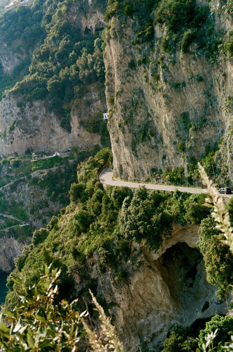 Costiera Amalfitana - Una strada stretta e tortuosa si snoda lungo ripide scogliere rocciose ricoperte di verde nella Costiera Amalfitana, con una grande grotta visibile nella parete rocciosa e un fogliame lussureggiante in primo piano: un vero e proprio scenario da Dolce Vita.