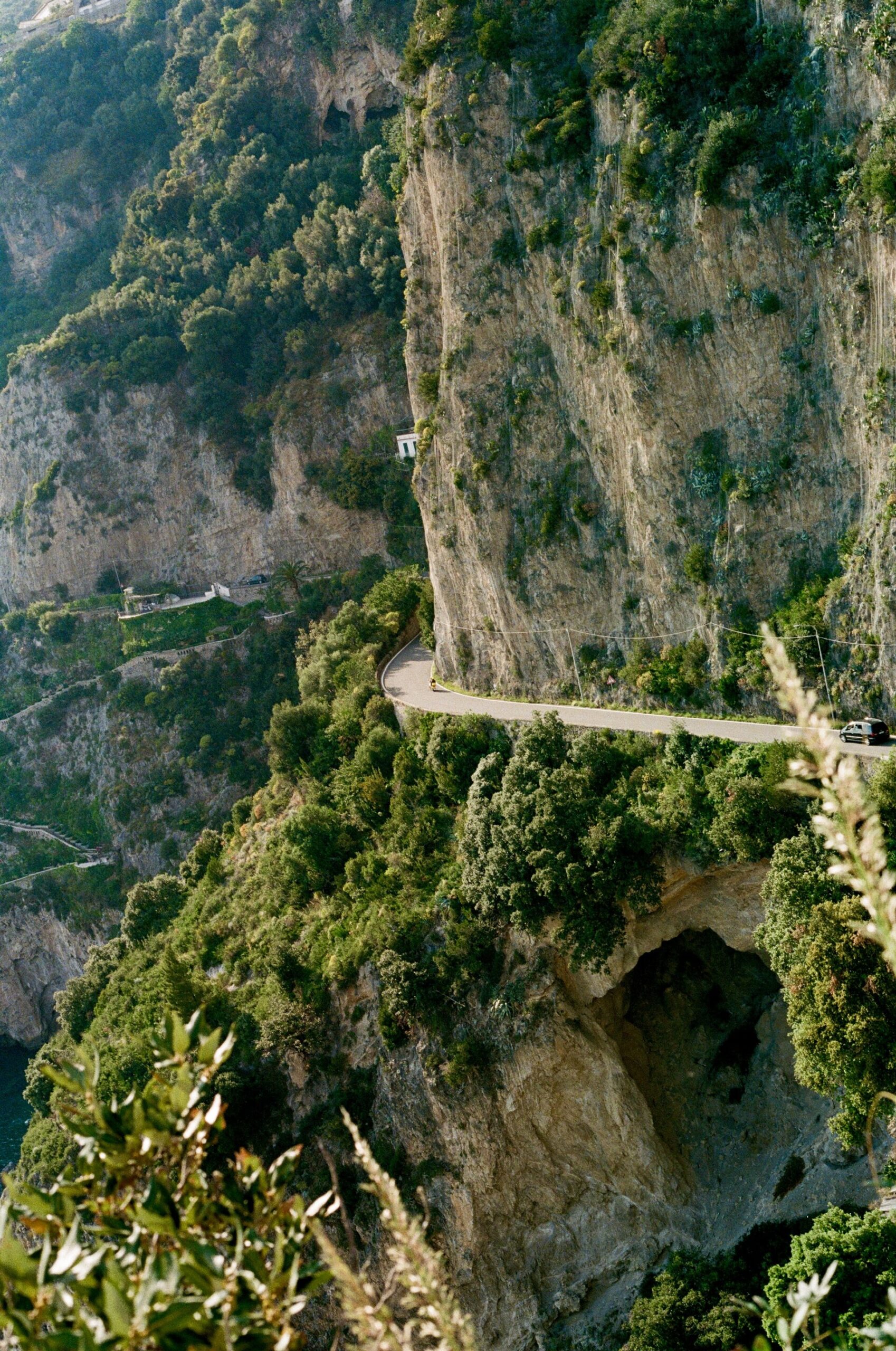 Costiera Amalfitana - Una strada stretta e tortuosa si snoda lungo ripide scogliere rocciose ricoperte di verde nella Costiera Amalfitana, con una grande grotta visibile nella parete rocciosa e un fogliame lussureggiante in primo piano: un vero e proprio scenario da Dolce Vita.