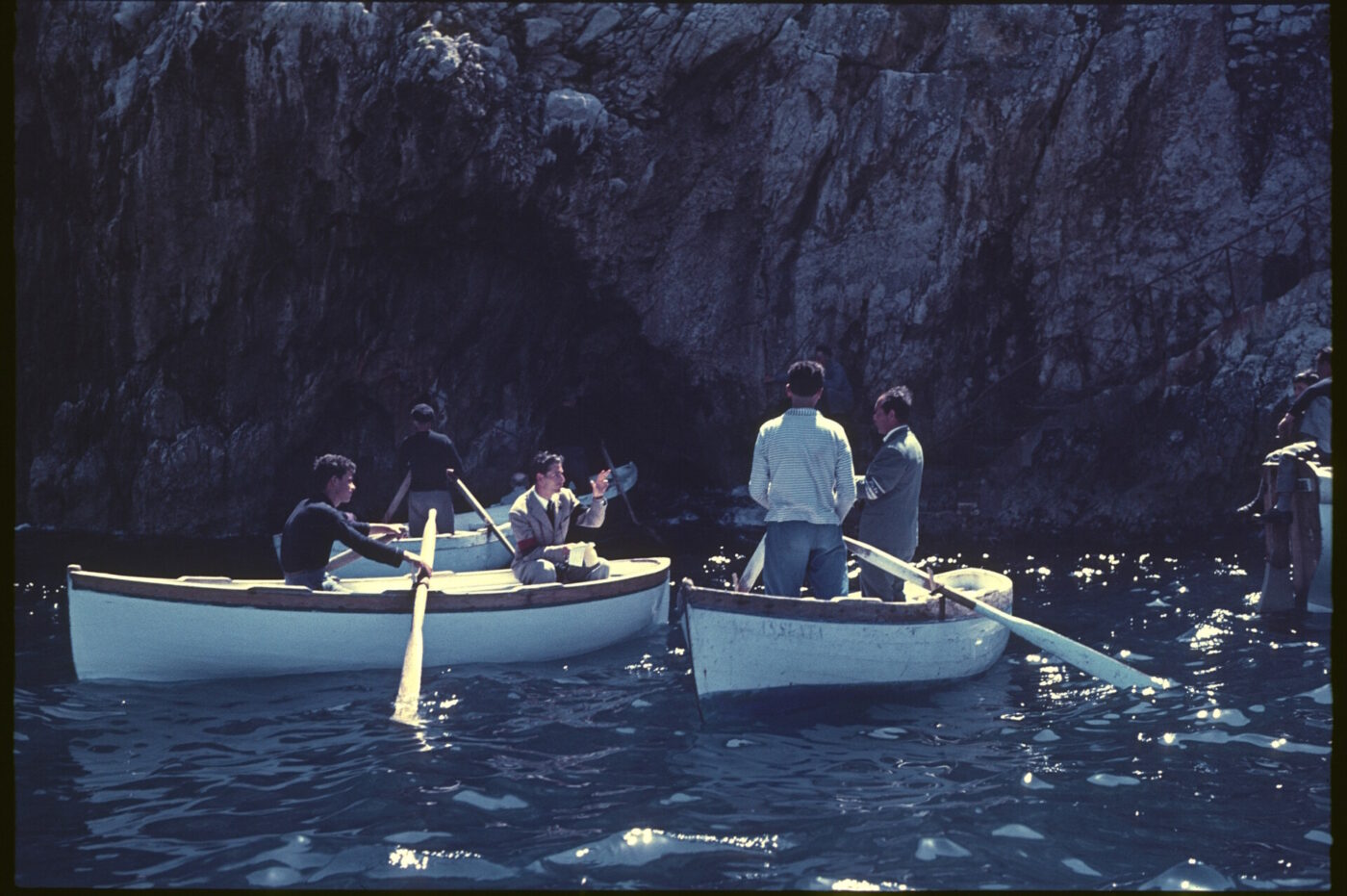 Barche a remi della Costiera Amalfitana - Tre barche a remi in legno con persone a bordo galleggiano vicino a una grotta rocciosa lungo la Costiera Amalfitana. Alcuni remano e altri parlano, godendosi i momenti di Dolce Vita mentre la luce del sole danza sull'acqua calma e blu.