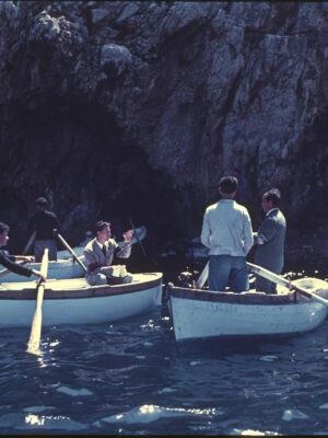 Four men in two small rowboats talk and gesture to each other on the calm blue water near a rocky cliff, basking in sunlight. Their boats float close together, savoring a dolce vita moment reminiscent of an Amalfi Coast holiday.