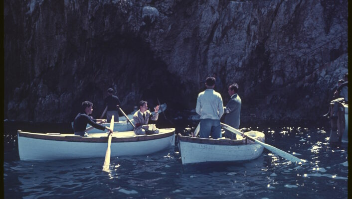 Four men in two small rowboats talk and gesture to each other on the calm blue water near a rocky cliff, basking in sunlight. Their boats float close together, savoring a dolce vita moment reminiscent of an Amalfi Coast holiday.