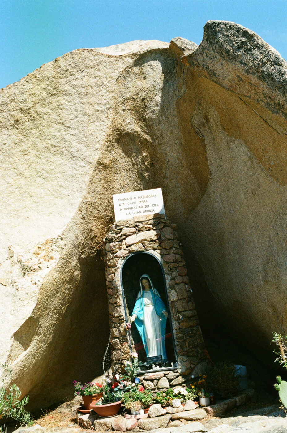 Statue of the Virgin Mary in Emerald Coast Sardinia - A statue of the Virgin Mary stands in a stone grotto at the base of a large rock, surrounded by potted plants, under a blue sky along the picturesque Emerald Coast Sardinia. A sign in Italian is displayed above the statue.