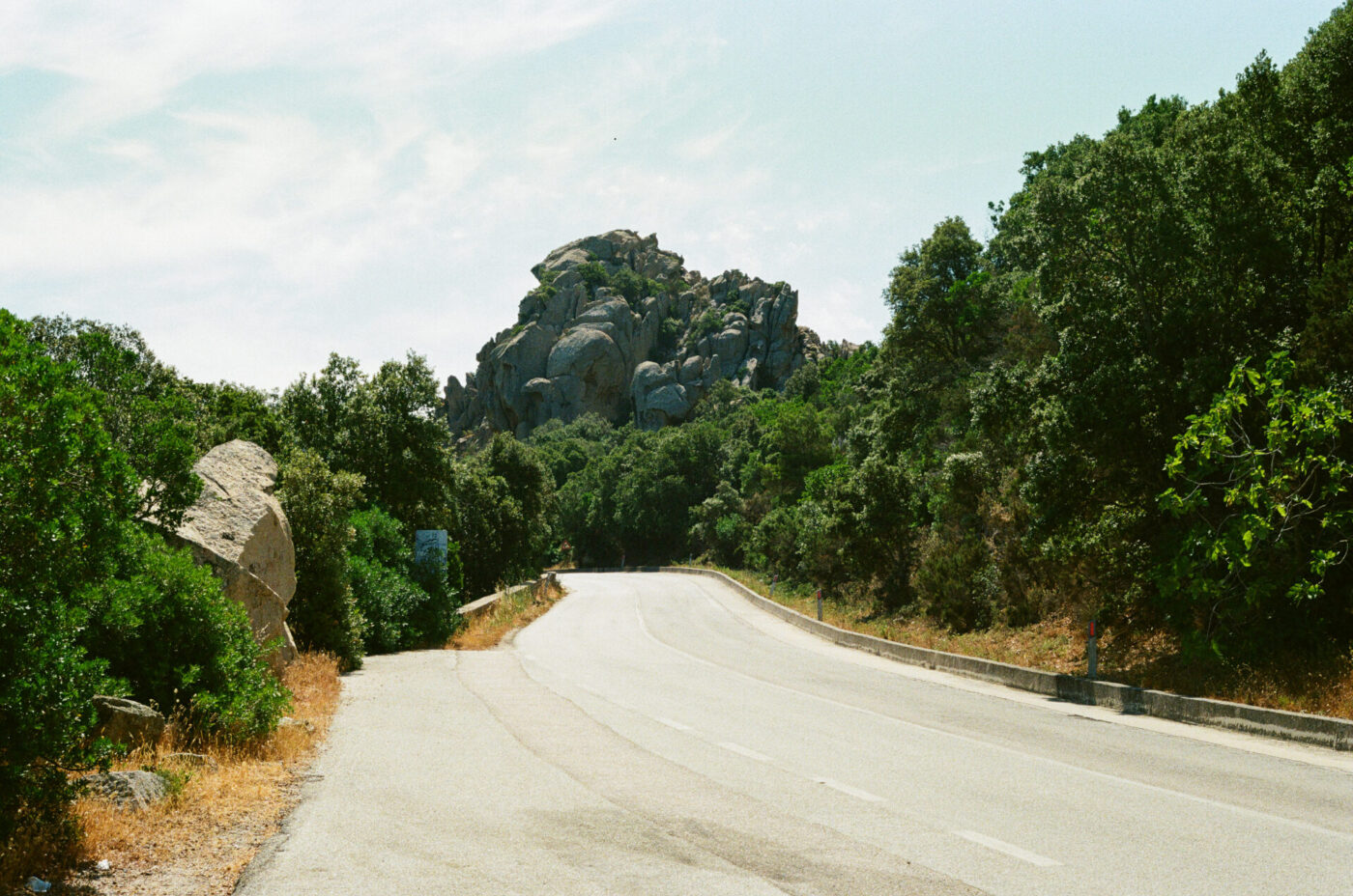 Emerald Coast Road - A curving empty road passes through lush green trees along the stunning Emerald Coast, leading toward a rocky hill under Sardinias bright, partly cloudy sky.