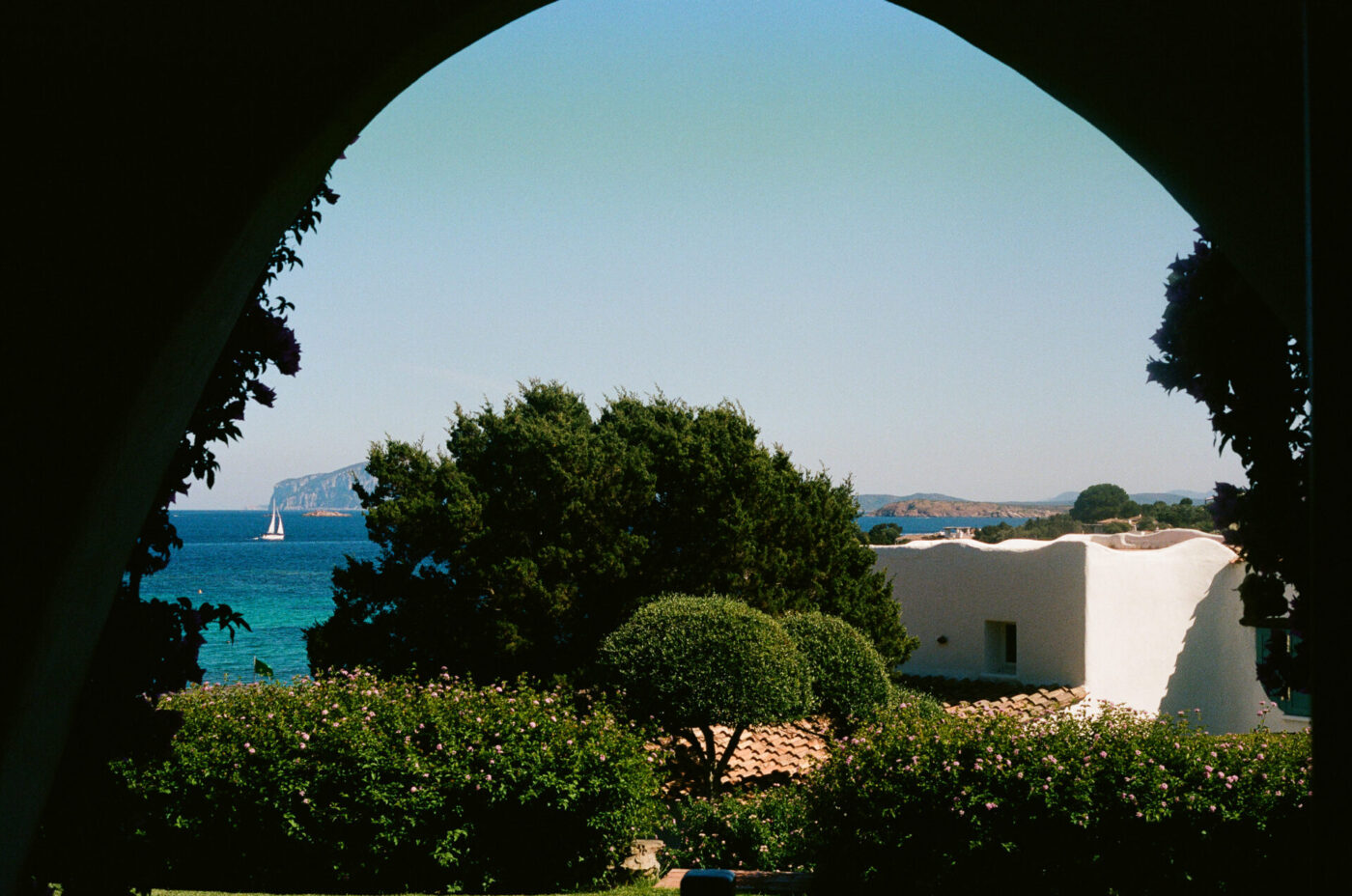 Emerald Coast View - View of the emerald coast Sardinia and a distant sailboat framed by an arched window, with trees, flowering bushes, and a white stucco building in the foreground under a clear blue sky.