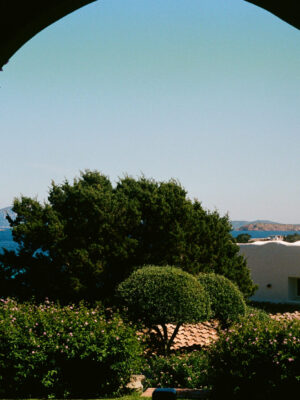 Emerald Coast View - View of the emerald coast Sardinia and a distant sailboat framed by an arched window, with trees, flowering bushes, and a white stucco building in the foreground under a clear blue sky.
