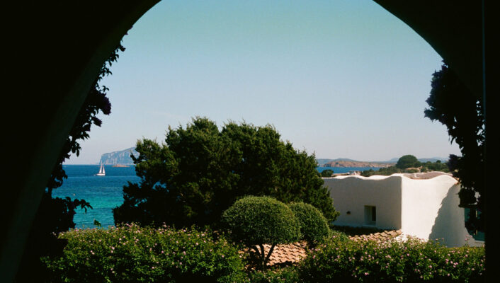 Vista attraverso un arco di un giardino lussureggiante, una casa bianca in stile mediterraneo, un mare blu, una barca a vela lontana e un'isola rocciosa sotto un cielo limpido in Costa Smeralda, Sardegna.