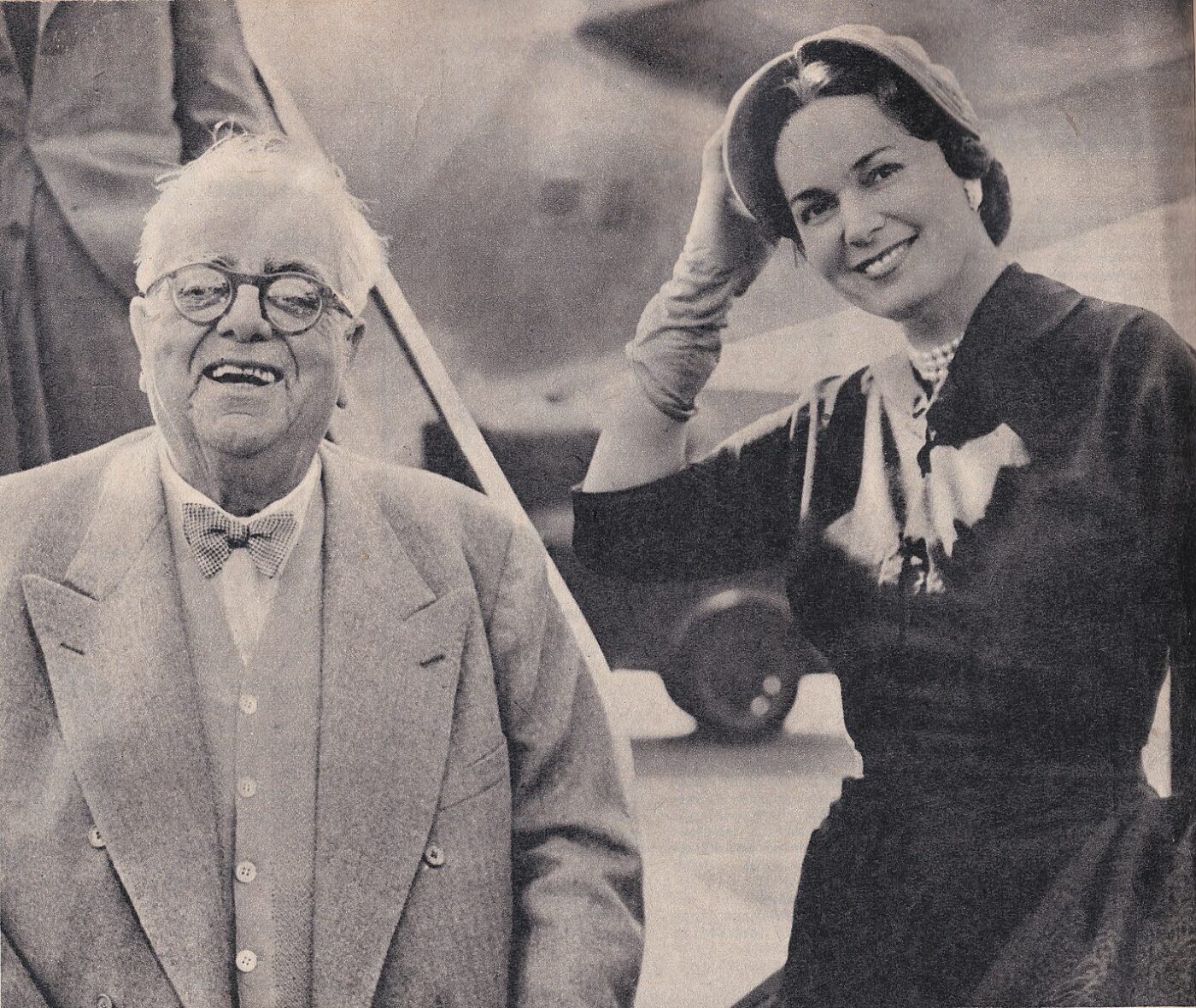 Aga_Khan_and_Begum_Yvette - An older man in a suit and bow tie smiles next to a young woman in a dress, gloves, and hat—touching her hat as they smile together. They stand outdoors along the Emerald Coast Sardinia, with an airplane visible in the background. If you provide a different or specific title, substitute accordingly.
