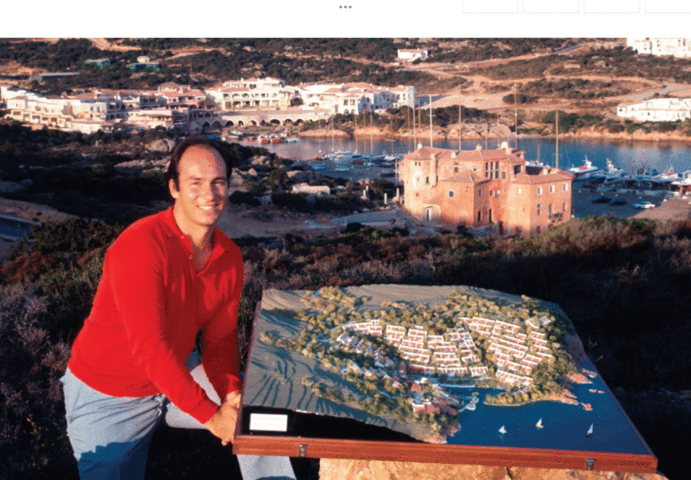 A man in a red shirt stands outdoors holding an architectural model of an Emerald Coast Sardinia resort with waterfront and hills.