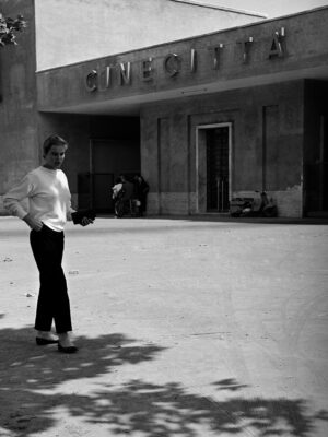 Cinecittà Film Studio - Una persona in top bianco e pantaloni scuri cammina all'esterno dello studio cinematografico di Cinecittà alla luce del giorno, con ombre e una facciata minimalista sullo sfondo.