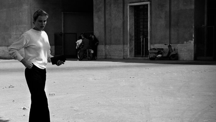 Cinecittà studios entrance - A woman in a white top and dark pants walks outside the entrance of Cinecittà studios, with the building’s sign visible above the doorway on a sunny day.