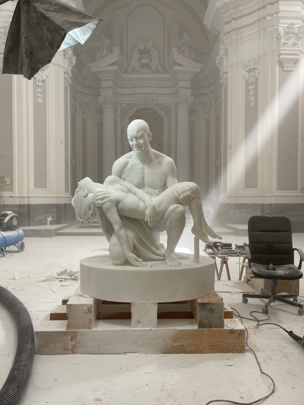 White sculpture of bald men down on his knees holding a woman's body like Michelangelo’s Pietà in a church studio with antique background in Napoli Campania in Italy