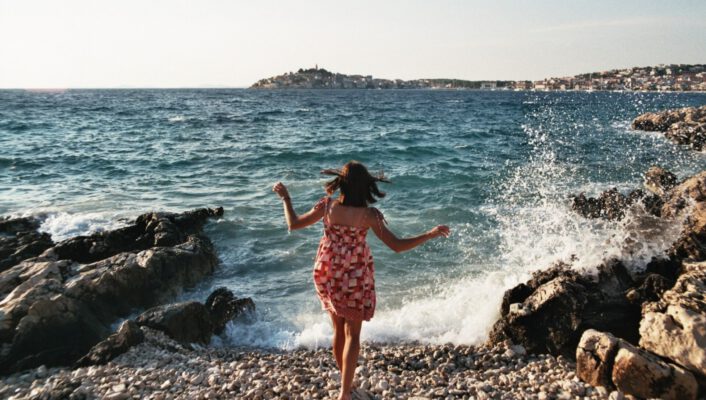 A young girl running into the sea as a wave splashes over a rocks beach and hills background in Italy