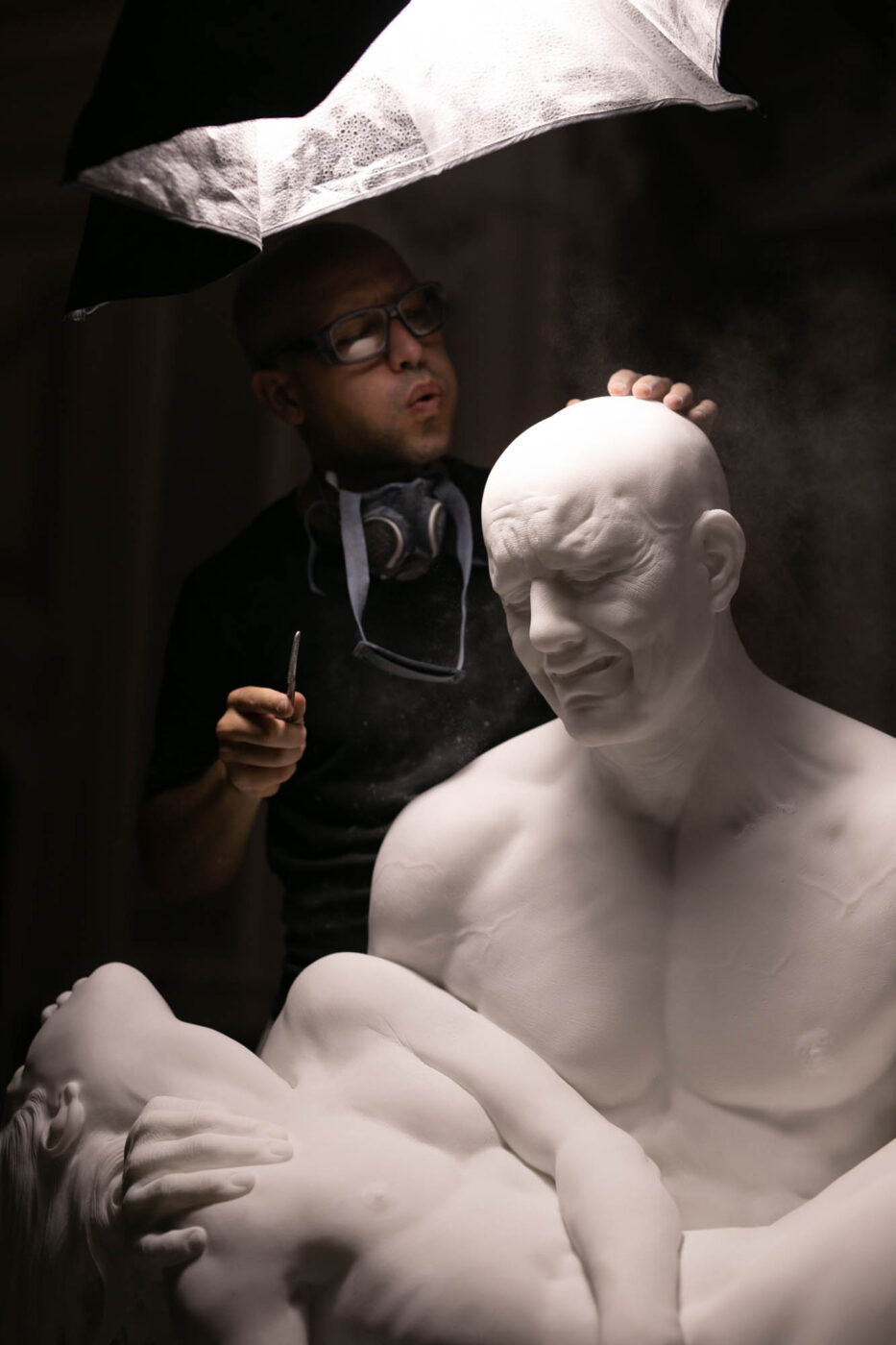 White sculpture of bald men down on his knees holding a woman's body like Michelangelo’s Pietà in a church studio with Jago artist wearing glasses and blowing its head in Napoli Campania in Italy