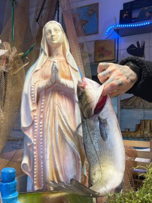 Tattooed men having a fish next to a virgin sculpture with fish nets behind and kitchen stuff Pescheria Mattiucci restaurant in Naples Campania in Italy