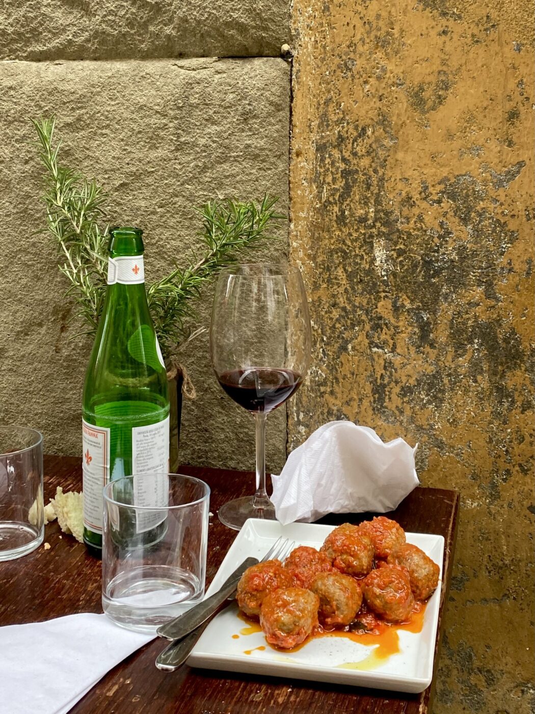 A plate of meatballs in tomato sauce sits on a wooden table beside an empty wine glass, a green glass bottle, and two empty water glasses, with a sprig of rosemary and textured yellow stone wall in the background.