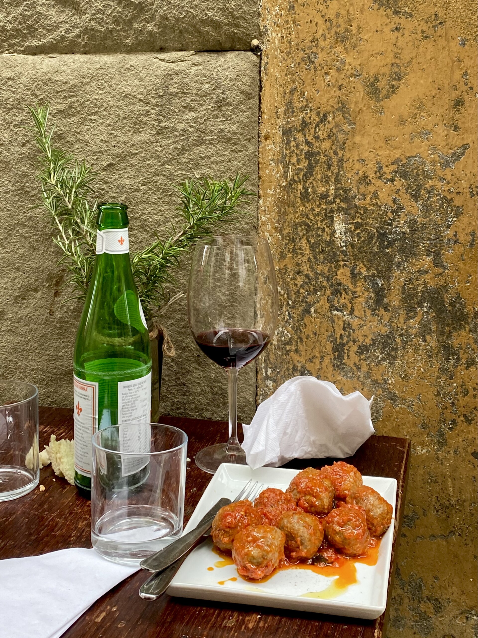 A plate of meatballs in tomato sauce sits on a wooden table beside an empty wine glass, a green glass bottle, and two empty water glasses, with a sprig of rosemary and textured yellow stone wall in the background.