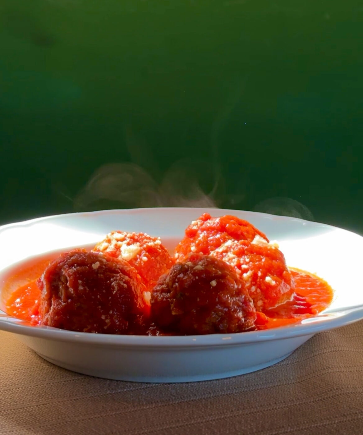 Five Steaming Meatballs - Five steaming meatballs topped with tomato sauce and grated cheese are served in a white bowl against a green background.