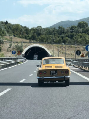Antique yellow fiat car driving in a highway heading to a tunnel surrounded by trees and mountains in the marche region of Italy