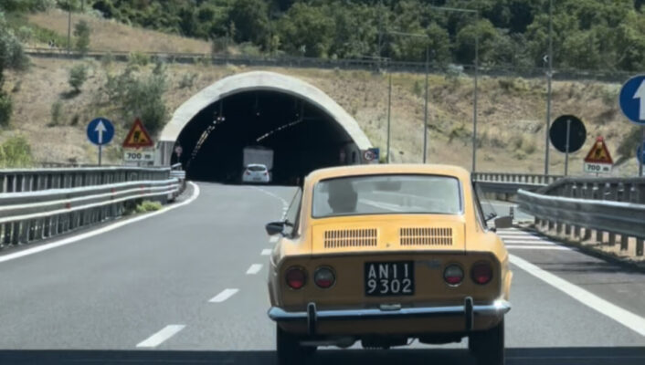 Antique yellow fiat car driving in a highway heading to a tunnel surrounded by trees and mountains in the marche region of Italy