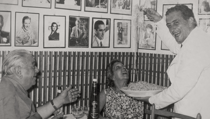 Vintage picture of elder people watching a chef serving fettuccine alfredo in typical restaurant seated in wooden table surrounded by frames in black and white in Rome Lazio in Italy
