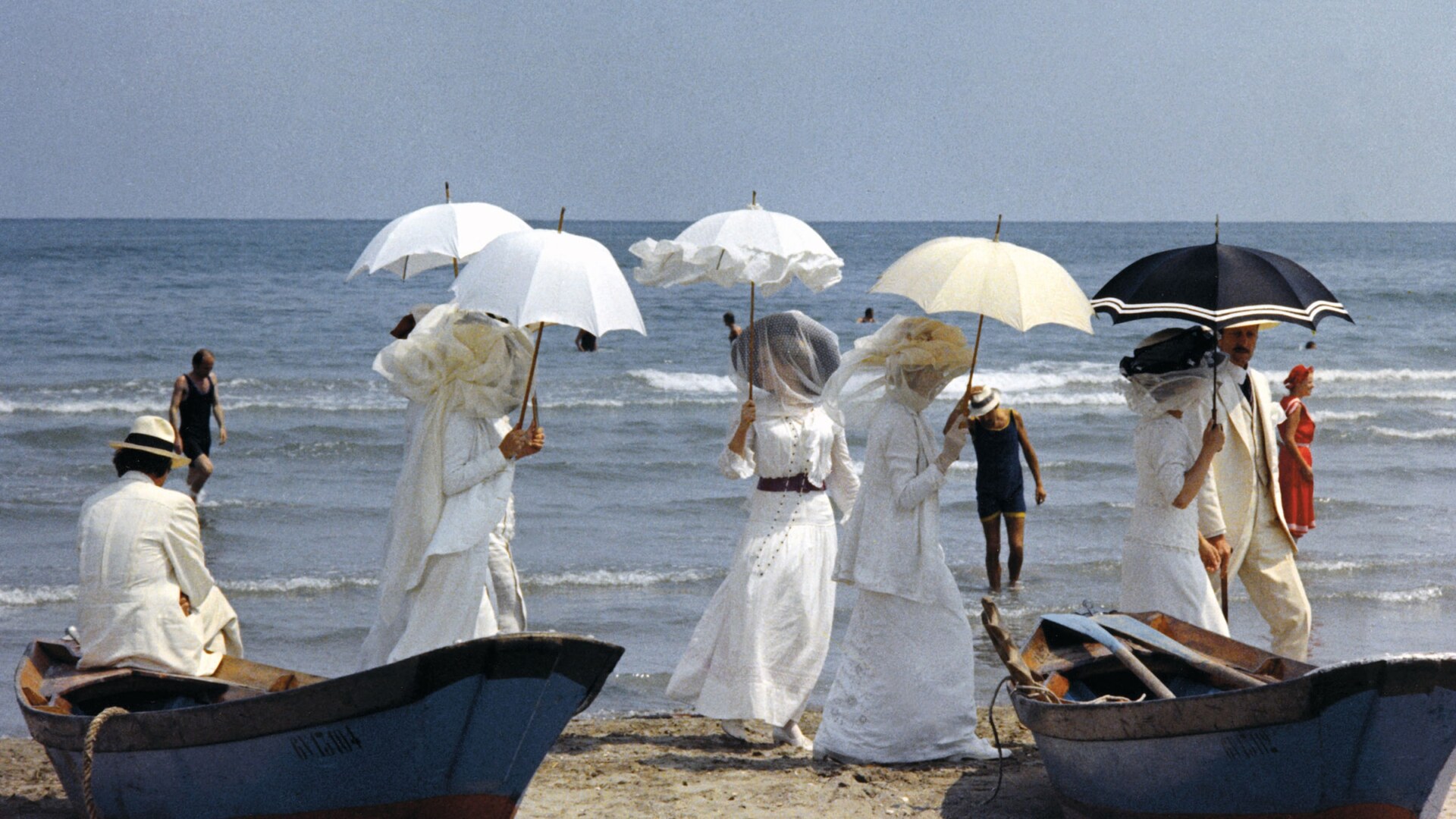 5 women dressed in white with veils and umbrellas walk along the beach near rowboats from the movie Death in Venice