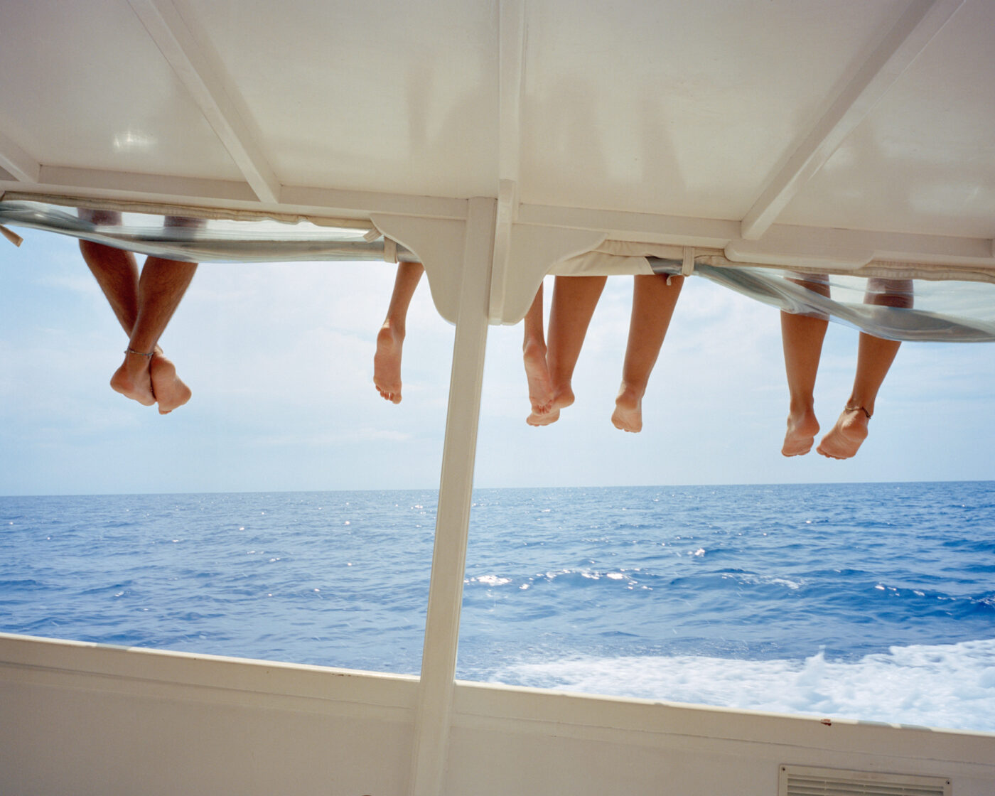photo by Michele Iacobini of four dangling pairs of legs over the side of the roof of a boat on the blue water