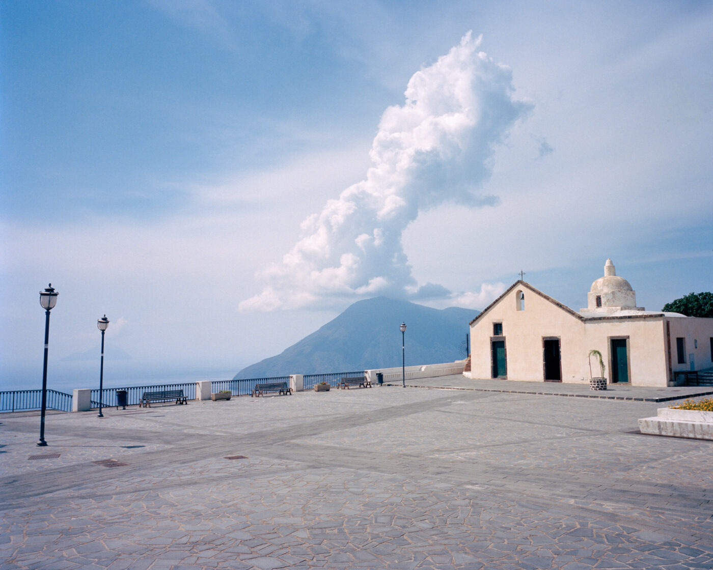 photo by Michele Iacobini of a simple white church on a hill overlooking the sea and a mountain and white fluffy cloud in the background