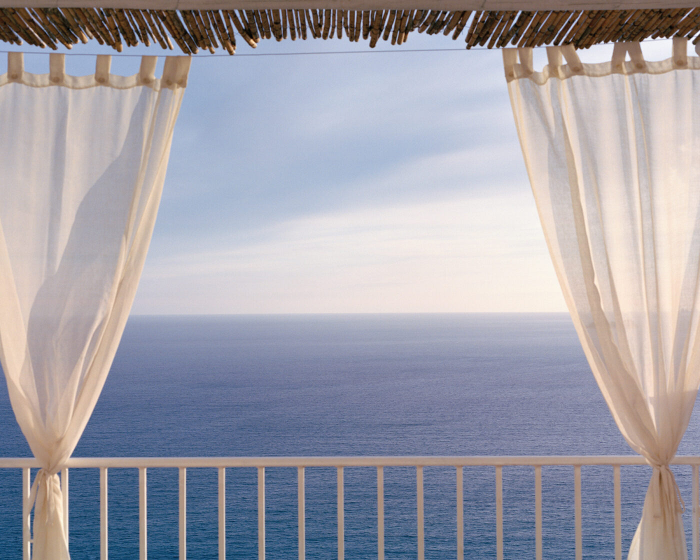 photo of the sea by Michele Iacobini looking out over a white railing balcony with two white curtains pulled to the side
