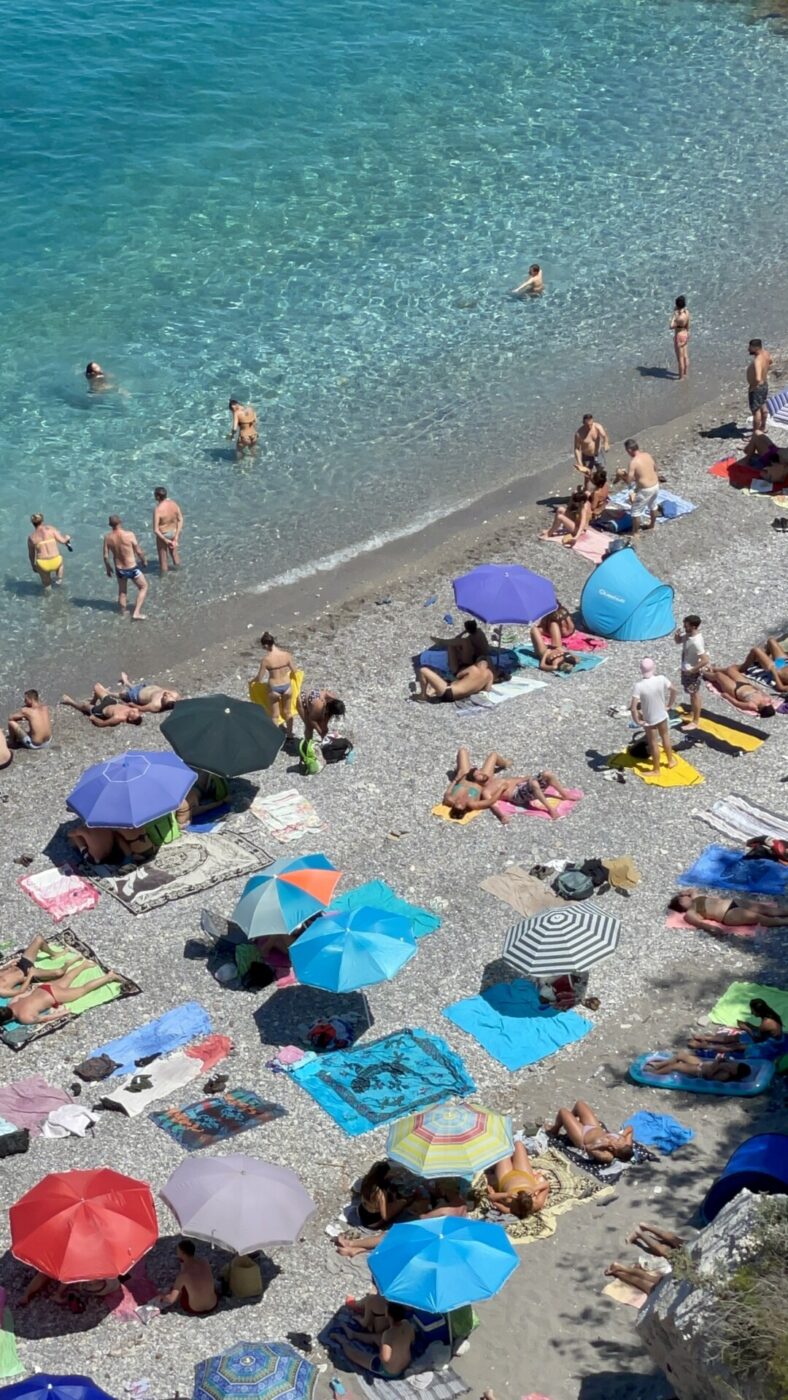 an overhead photo of many brightly colored umbrellas and people tanning on the sandy beach with clear blue water