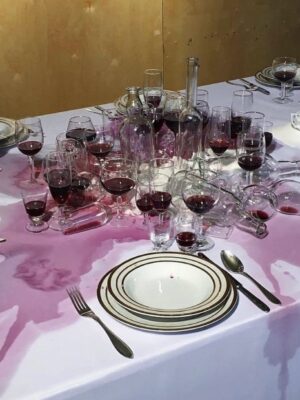 red wine spilled all over a white tablecloth on a nicely set table with nearly 20 wine glasses collected in the center