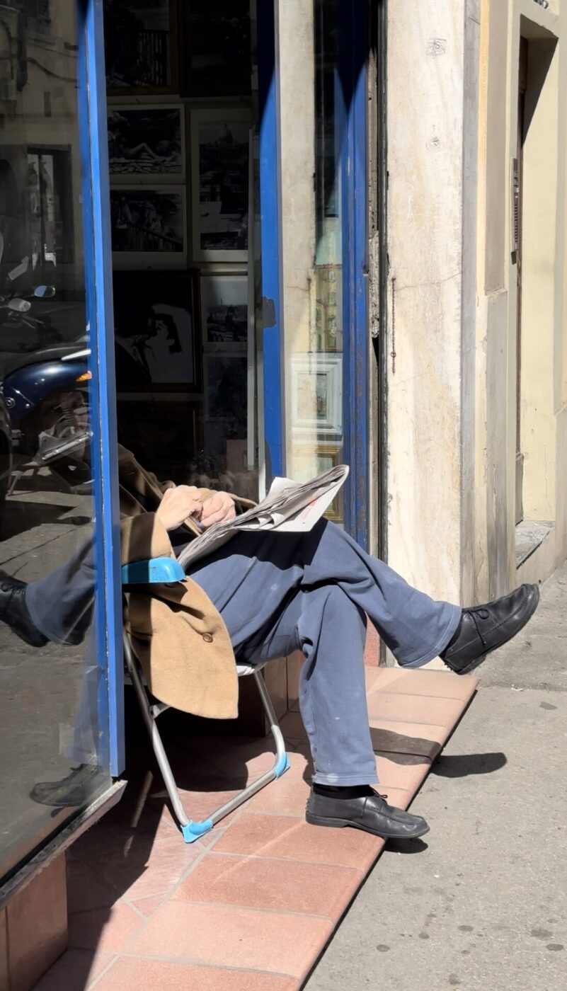a man sitting in a storefront with the lower half of his body sticking out into the sun